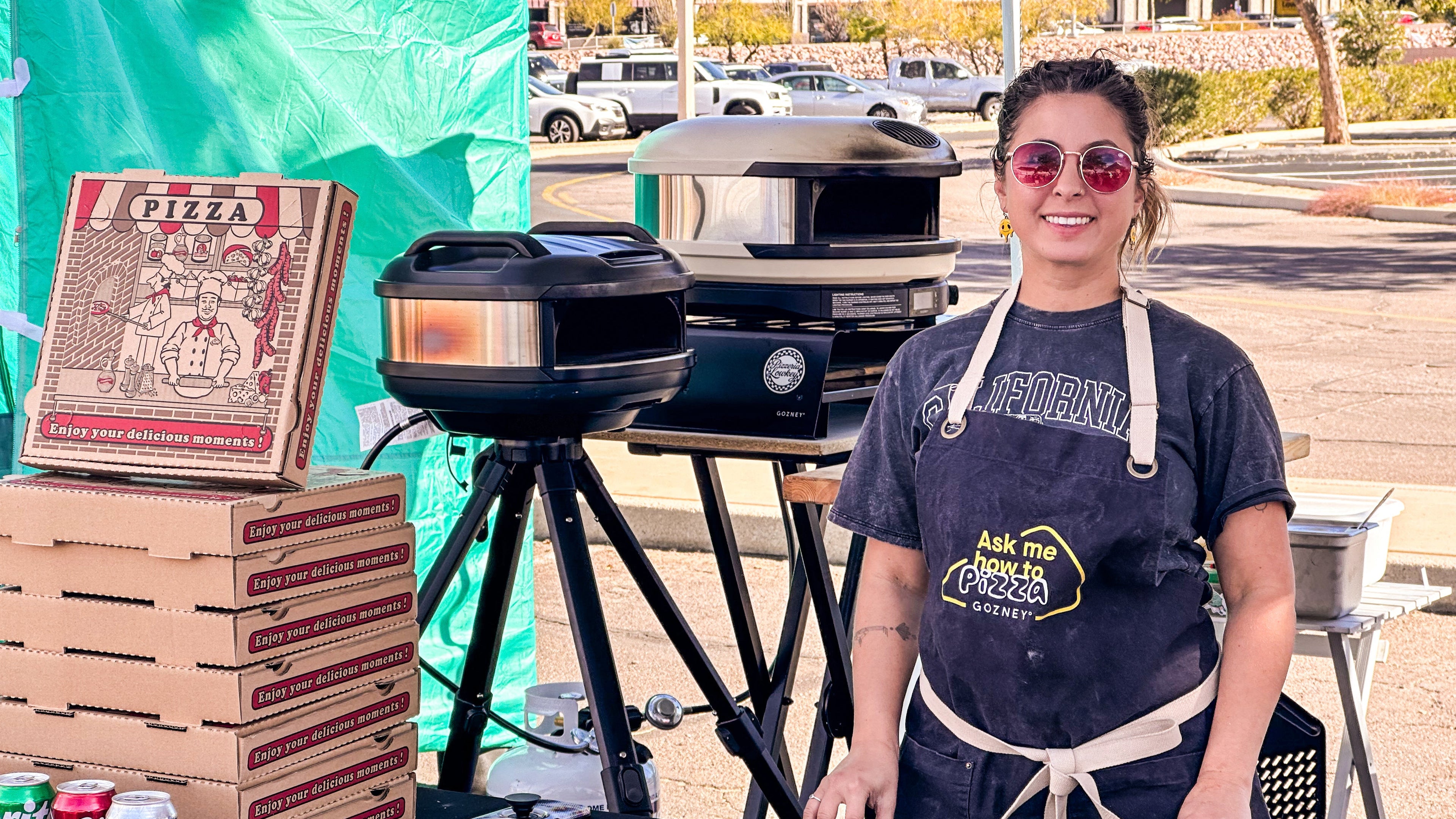 Person in a pizza-themed apron standing behind a table with pizza boxes and a small oven.